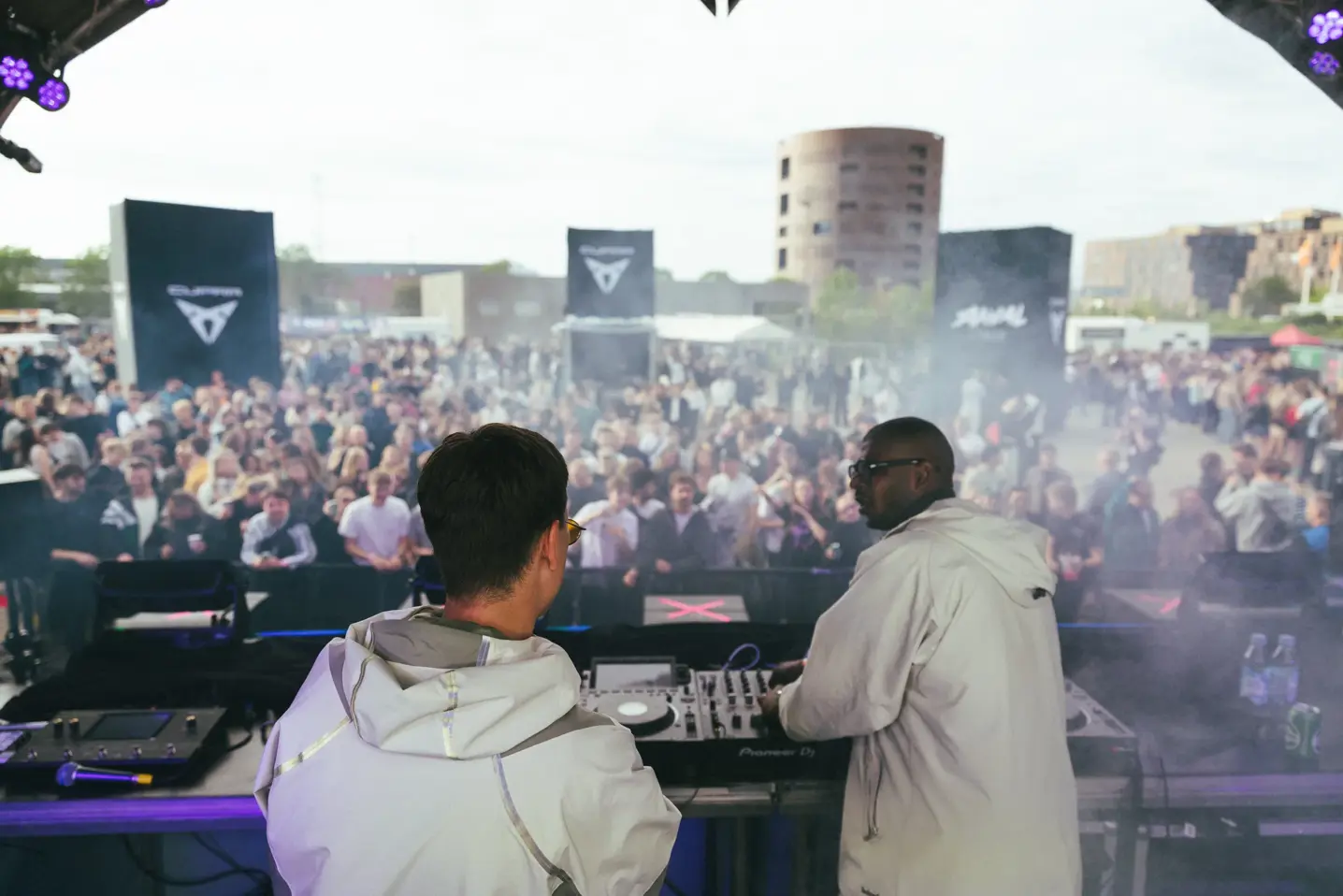 To DJs spiller musik på en udendørs scene foran en stor, tæt pakket festivalpublikum.