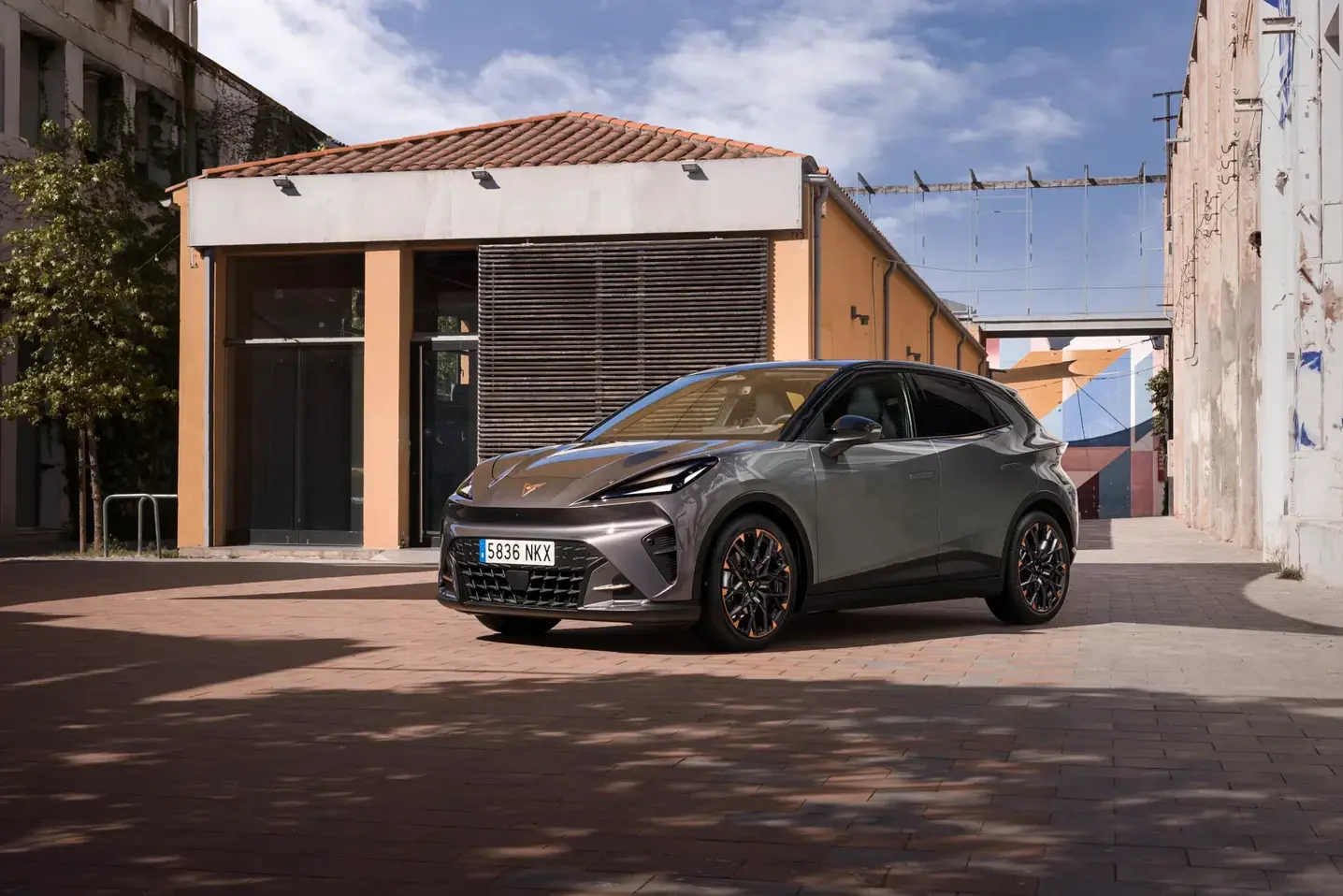 En grå CUPRA Raval elbil holder parkeret på en solrig brostensplads mellem moderne bygninger.