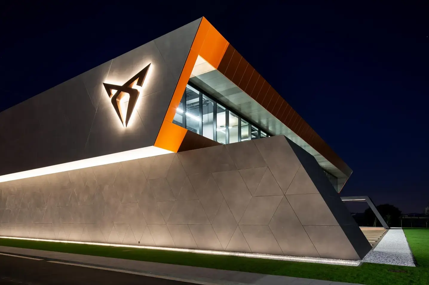 Moderne, skarpt vinklet CUPRA-bygning oplyst om natten med stort lysende logo på facaden.