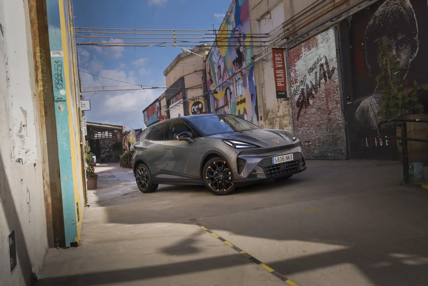 En grå CUPRA Raval elbil holder parkeret i en farverig, graffitifyldt bygyde under blå himmel.