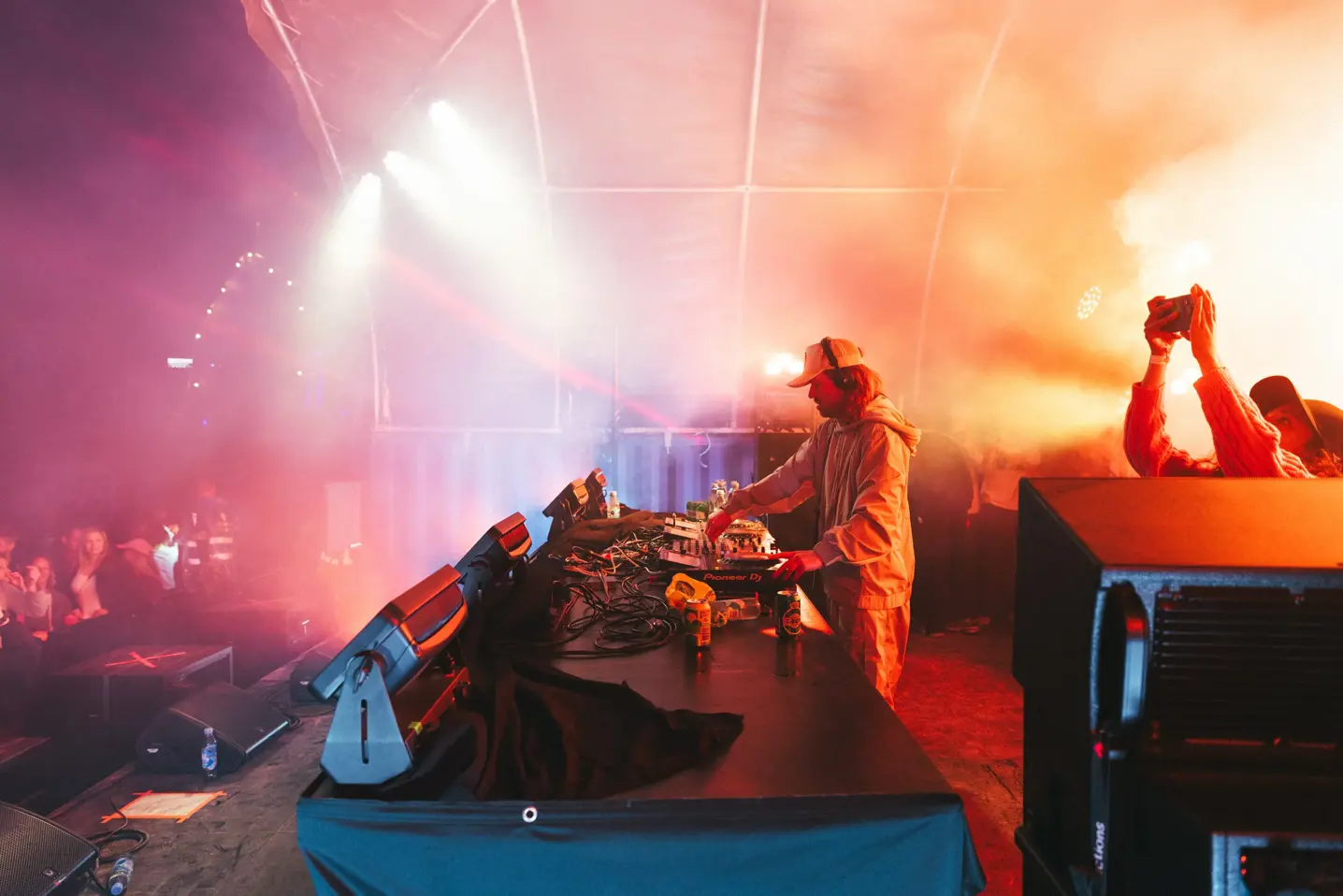 DJ optræder på en farverig, røgfyldt festivalscene, mens publikum danser og filmer foran scenen.