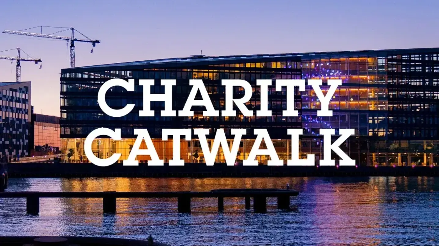 Aftenbillede af en moderne havnefrontbygning med teksten "Charity Catwalk" hen over vandet.
