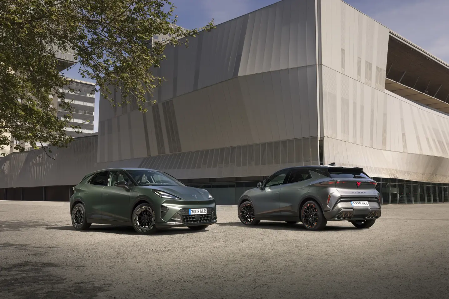 To CUPRA Raval elbiler står parkeret foran en stor, moderne bygning i beton og glas.